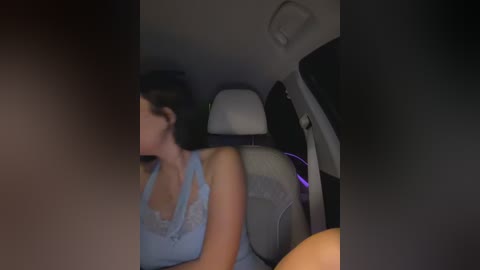 Video of a woman in a car, seen from the front passenger seat, wearing a light blue sleeveless top, her back slightly arched, revealing her buttocks. The car interior is dimly lit.