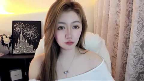 Video of a young Asian woman with long, straight, light brown hair, wearing a white off-shoulder top, standing indoors. Background includes a black-framed cityscape print, lace curtains, and a vase with white flowers.