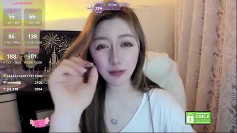 A video of a young Asian woman with long brown hair, wearing a white top, sitting indoors, holding a smartphone displaying a \"Live\" app. The room has beige curtains and a white couch.