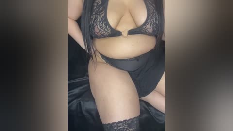 Video of a voluptuous woman in black lace lingerie, including a sheer bra and high-waisted panties, with long black hair cascading over her shoulders, sitting on a black satin surface.
