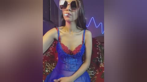 Video of a young woman with fair skin, long brown hair, and large sunglasses, wearing a sheer blue babydoll lingerie with red lace and a red bow, seated on a floral-patterned red couch.