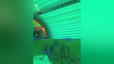 A video of a green-tinted tanning bed with a person's leg adorned with a blue ribbon and intricate tattoo. The tanning bed has a curved, metallic interior.