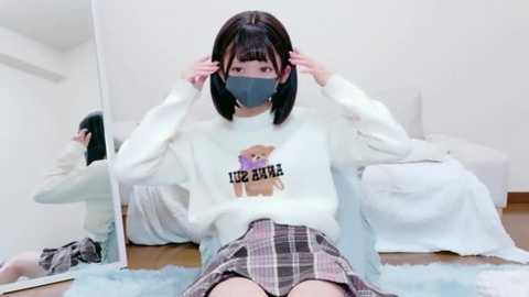 Video of an Asian woman with straight black hair, wearing a white sweatshirt with a bear graphic, a face mask, and a plaid skirt, adjusting her mask in a minimalist white room.