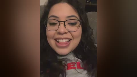 Video of a smiling young woman with curly dark hair, wearing glasses, a gray t-shirt with red letters, and a necklace, indoors.