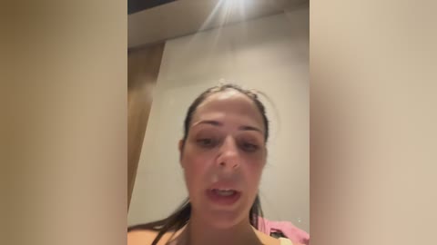 Media: A blurred video of a young woman with light skin and brown hair, wearing a pink top, taken indoors with a bright ceiling light in the background.