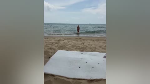 Video of a sandy beach with a white, cushioned lounger in the foreground, a lone person in a red swimsuit standing in the shallow ocean water in the middle background, and a partly cloudy sky overhead.