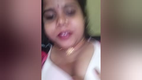 A blurred video of a South Asian woman with dark hair, wearing a white top and gold necklace, looking distressed.