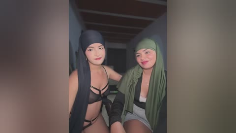 Video of two women wearing black and green headscarves, black lingerie, and green jackets, standing in a dimly lit hallway with wooden ceiling beams.