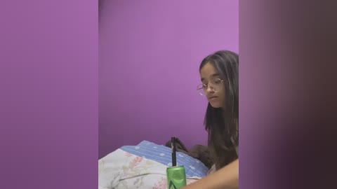 Video of a young woman with long brown hair, wearing glasses, holding a green umbrella against a purple wall. She has a neutral expression.