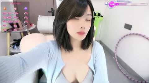 A video of an East Asian woman with shoulder-length black hair, fair skin, and a slender physique, wearing a light blue cardigan, partially revealing a black bra. She sits on a white couch in a modern living room with a pink hula hoop nearby.