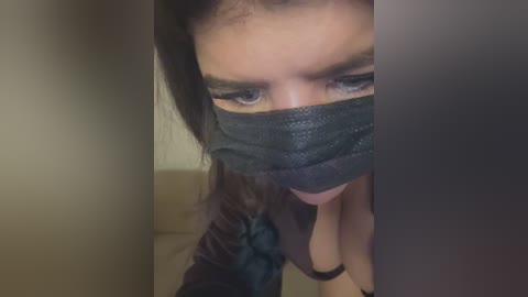Video of a woman with long dark hair, wearing a black face mask, showing only her eyes and mouth. She is topless, revealing cleavage.