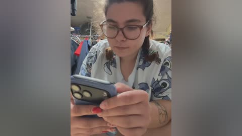 Video of a young woman with glasses, dark hair tied back, wearing a patterned shirt, intensely focused on a blue flip phone. She has red-painted nails and a tattoo on her wrist. Background includes clothing on racks and a blurred beige wall.