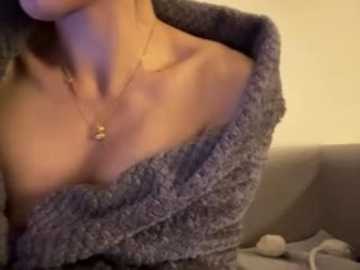 A video of a woman's partially exposed chest, wearing a grey, off-the-shoulder sweater, a delicate gold necklace, and a blurred beige couch background.