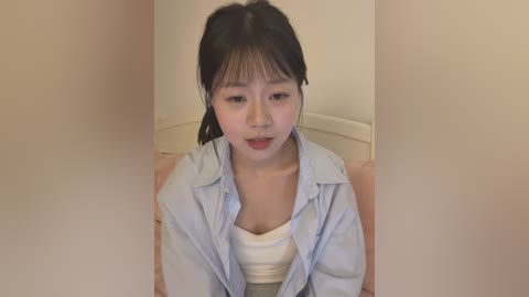 Video of an Asian woman with fair skin, dark hair in a ponytail, wearing a light blue shirt over a white tank top, sitting indoors with a neutral background.