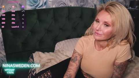 A video of a blonde woman with tattoos, wearing a beige top, seated on a bed with a green tufted headboard. The background features a floral-patterned wallpaper.