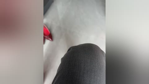 Video of a close-up of a person's shoulder and upper back, with a red object (possibly a hair clip or accessory) visible on the left side. The background is blurred, showing a neutral-colored wall. The subject appears to be wearing a dark grey shirt.