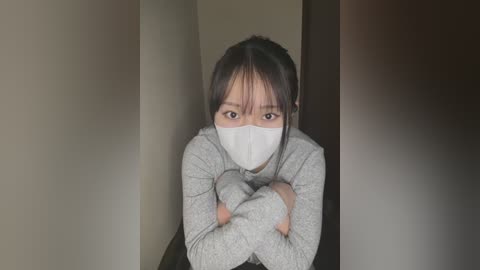 A video of a young Asian woman with medium skin tone, wearing a gray long-sleeved top, white surgical mask, and arms crossed, standing in a dimly lit hallway.