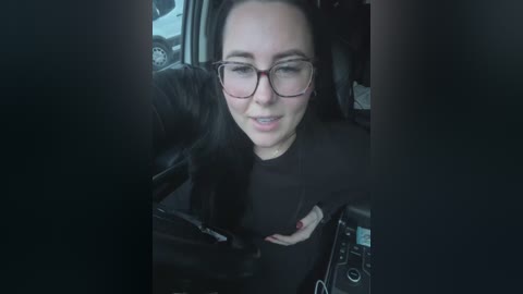 Video of a smiling woman with glasses, black hair, and a black jacket, driving a car with a black interior, showing a smartphone on the dashboard.