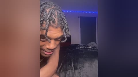 Video of a Black man with medium skin tone and dreadlocks, lying on a bed in a dimly lit room with blue LED lights, wearing a black shirt.