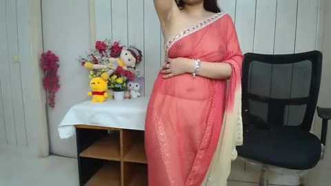 Video of a woman in a semi-transparent red saree, standing in front of a white wall with floral decorations, a chair, and a yellow stuffed toy.