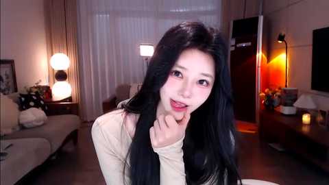 Media: Video of an East Asian woman with long black hair, light skin, and red lipstick, wearing a beige top, smiling in a warmly lit living room with a couch, lamp, and modern decor.