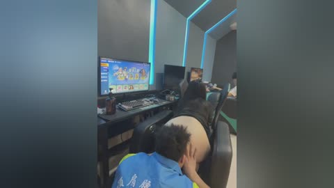 Video of an Asian man in a gaming room, slumped over a desk with a blue gaming chair, playing a video game with a large monitor showing a colorful game interface.