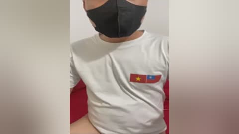 A video of a person wearing a white T-shirt with a red and blue Vietnamese flag patch on the left chest, black face mask, and black hair, partially visible against a white and red background.