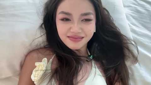 Media: Video of a young woman with long dark hair, wearing white headphones, lying on a white bed, smiling softly, with a relaxed, casual vibe.