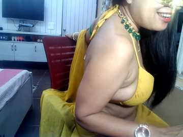 Video of a woman in a mustard yellow saree with a green beaded necklace, sitting in a bedroom with a red chair and a TV on a white cabinet.