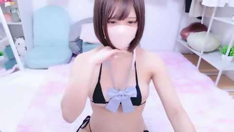 A video of an Asian woman with short brown hair, wearing a revealing black bikini with a blue bow, standing in a pink-carpeted room with light furniture.