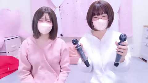 Video of two young Asian women with short brown hair, wearing pink robes, and surgical masks, holding microphones, in a pink-themed room with soft furnishings.