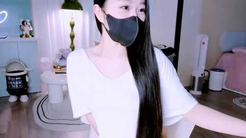Video of a young Asian woman with long black hair, wearing a black face mask, white T-shirt, and standing indoors with a white toilet, green lamp, and pink bed in the background.