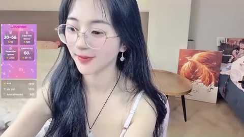 Media: Video of an East Asian woman with long black hair, glasses, and a white tank top, smiling in a bedroom with a bed, table, and artwork.