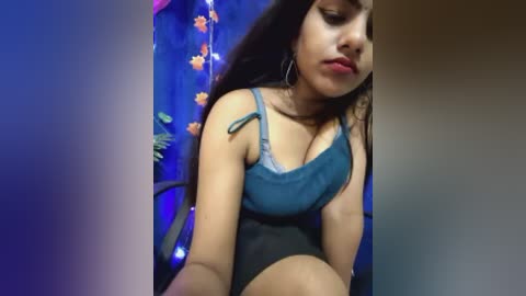A video of a young South Asian woman with long black hair, medium skin tone, and medium build, wearing a blue tank top and black shorts, sitting in a dimly lit room with blue and orange floral wallpaper.