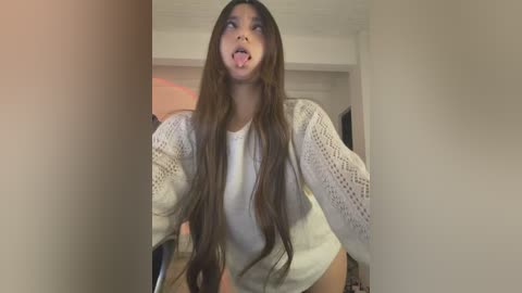 A video of a young woman with long brown hair, wearing a white crochet cardigan, sticking out her tongue, taken indoors with blurred background.