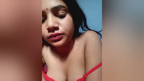 Video of a South Asian woman with medium-brown skin and long black hair, wearing a red lace bra. She has a nose ring and a slight smile, with a blurred background.