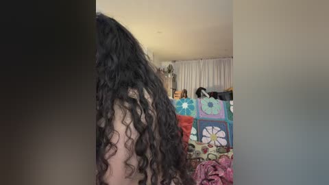 Video of a curly-haired person with long black hair, viewed from behind, in a cluttered, colorful living room with a patchwork quilt, white curtains, and assorted objects.