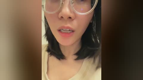 Media: Video of an Asian woman with light skin, short black hair, wearing clear-rimmed glasses, light pink lipstick, and a beige top. The background is blurred.