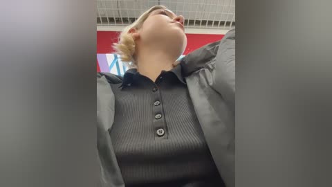 A video of a blonde woman in a gray pinstripe blazer and black ribbed shirt, captured from a low angle, with a red wall and white ceiling tiles in the background.
