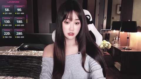 Video of an East Asian woman with long black hair, wearing a grey off-shoulder top, sitting on a gaming chair in a modern, dimly lit room.