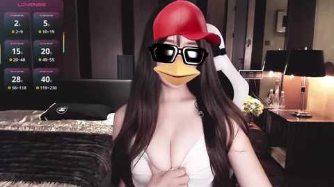 Video of a woman with long dark hair, wearing a red baseball cap, black sunglasses, and a white tank top, standing in a modern bedroom with a bed, nightstand, and TV displaying weather info.