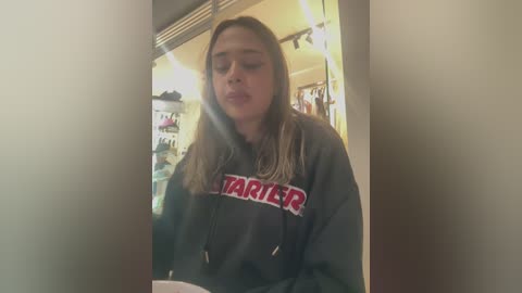 Video of a young woman with light brown hair wearing a gray Star Wars hoodie, standing in a dimly lit room with shelves and hanging clothes in the background.