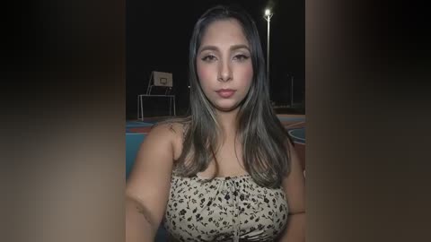 A video of a young woman with long, straight dark hair, fair skin, and a medium build, wearing a strapless, leopard-print dress. She stands in an outdoor setting with dim lighting, a basketball hoop, and a bench in the background.