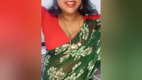 Media: Video of a South Asian woman with medium brown skin, wearing a green sari with white floral pattern, red blouse, gold necklace, and earrings. Background has a red, green, and white gradient.
