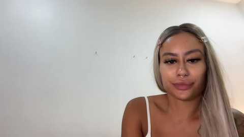 Video of a young woman with long, straight, platinum blonde hair, wearing a white spaghetti-strap tank top. She has light brown skin, a slender physique, and a small nose ring. Background is a plain, off-white wall.