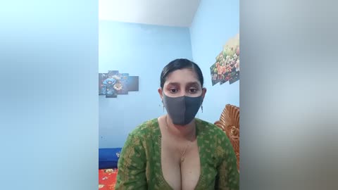 Video of a South Asian woman with medium skin tone, large breasts, wearing a green patterned blouse, black face mask, and dark hair, standing indoors against a light blue wall with floral decor.