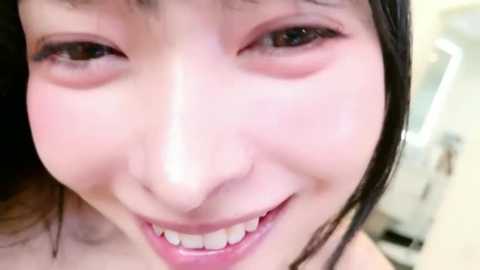 Close-up video of a smiling Asian woman with fair skin, brown eyes, and straight black hair. She has a light blush on her cheeks and wears minimal makeup. The background is blurry, suggesting an indoor setting.