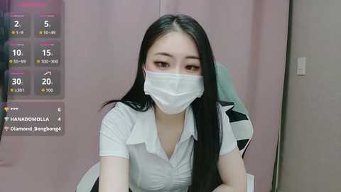 Video of an Asian woman with long black hair, wearing a white face mask and a white blouse, sitting in a chair in a pink room, with a digital display showing temperature and humidity readings.
