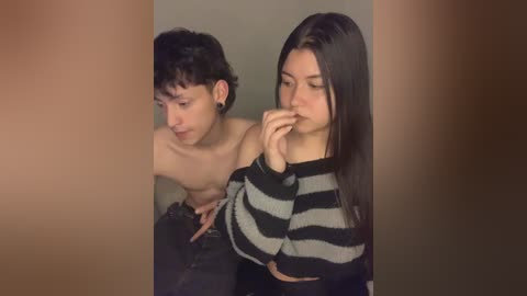 Media: Video of a shirtless young man with curly hair, wearing a black and white striped sweater, and a young woman with long dark hair, both looking at a camera through a blurry background.