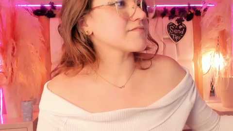 Media: Video of a young woman with light skin, brown hair, and glasses, wearing a white off-the-shoulder top, standing against a rustic, orange-painted wall with a heart-shaped \"Send It\" sign. Warm, ambient lighting.